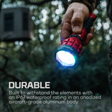 Illuminated red and blue flashlight held in a hand against a blurred outdoor background.