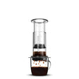 Aeropress coffee maker with a clear chamber and dark coffee dripping into a glass mug below.