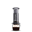 Cylindrical coffee press device with a clear chamber and black body, positioned over a glass mug containing brewed coffee.