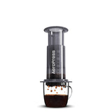 Cylindrical coffee press device with a clear chamber and black body, positioned over a glass mug containing brewed coffee.