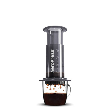 Cylindrical coffee press device with a clear chamber and black body, positioned over a glass mug containing brewed coffee.