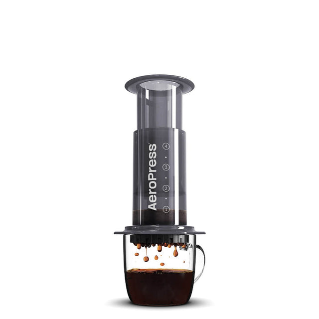 Cylindrical coffee press device with a clear chamber and black body, positioned over a glass mug containing brewed coffee.