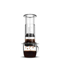 Aeropress coffee maker with a clear chamber and dark coffee dripping into a glass mug below.